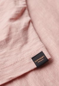 Lichtroze stof met een subtiele textuur, voorzien van een zwart label met de gouden opschrift "SUPERDRY". De stitching is netjes en discreet.