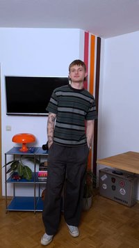 Νεαρός με τατουάζ που φοράει ριγέ πουκάμισο και σκούρο παντελόνι στέκεται σε δωμάτιο με τηλεόραση, πορτοκαλί φωτιστικό, ριγέ τοιχογραφία και μεταλλικό ράφι με φυτά.