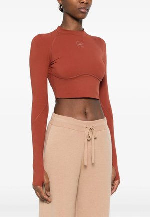 Donna che indossa un top corto a maniche lunghe color ruggine e pantaloni beige con coulisse, in piedi davanti a uno sfondo bianco uniforme.
