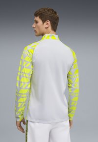 Långärmad idrottströja med vit kropp och neongul geometrisk mönster på ärmarna. Har ett lätt, fukttransporterande tyg.
