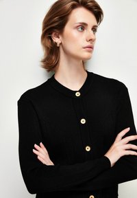 Svart ribbad cardigan med rund halsringning, prydd med två gyllene knappar framtill. Slät yta och långa ärmar.