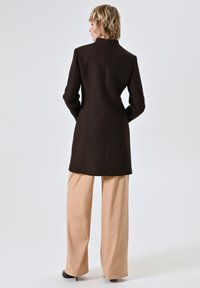 Vrouw met kort blond haar die zich omdraait, gekleed in een strakke donkerbruine jas, beige wijde broek en zwarte hakken, tegen een witte achtergrond.