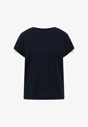 Femme souriante debout à l'intérieur, portant un haut bleu marine à manches courtes, un jean large bleu clair et des chaussures beiges sans lacets devant un mur beige.