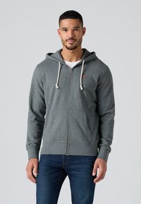 Felpa grigia con zip e cappuccio regolabile, polsini a costine, tasche frontali e una piccola etichetta rossa sul petto sinistro. Tessuto morbido e texturizzato.