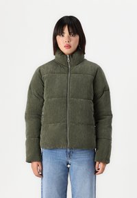 Groene corduroy pufferjack met een hoge kraag, quilted ontwerp, voorrits en twee zijzakken; gecombineerd met blauwe denim jeans.