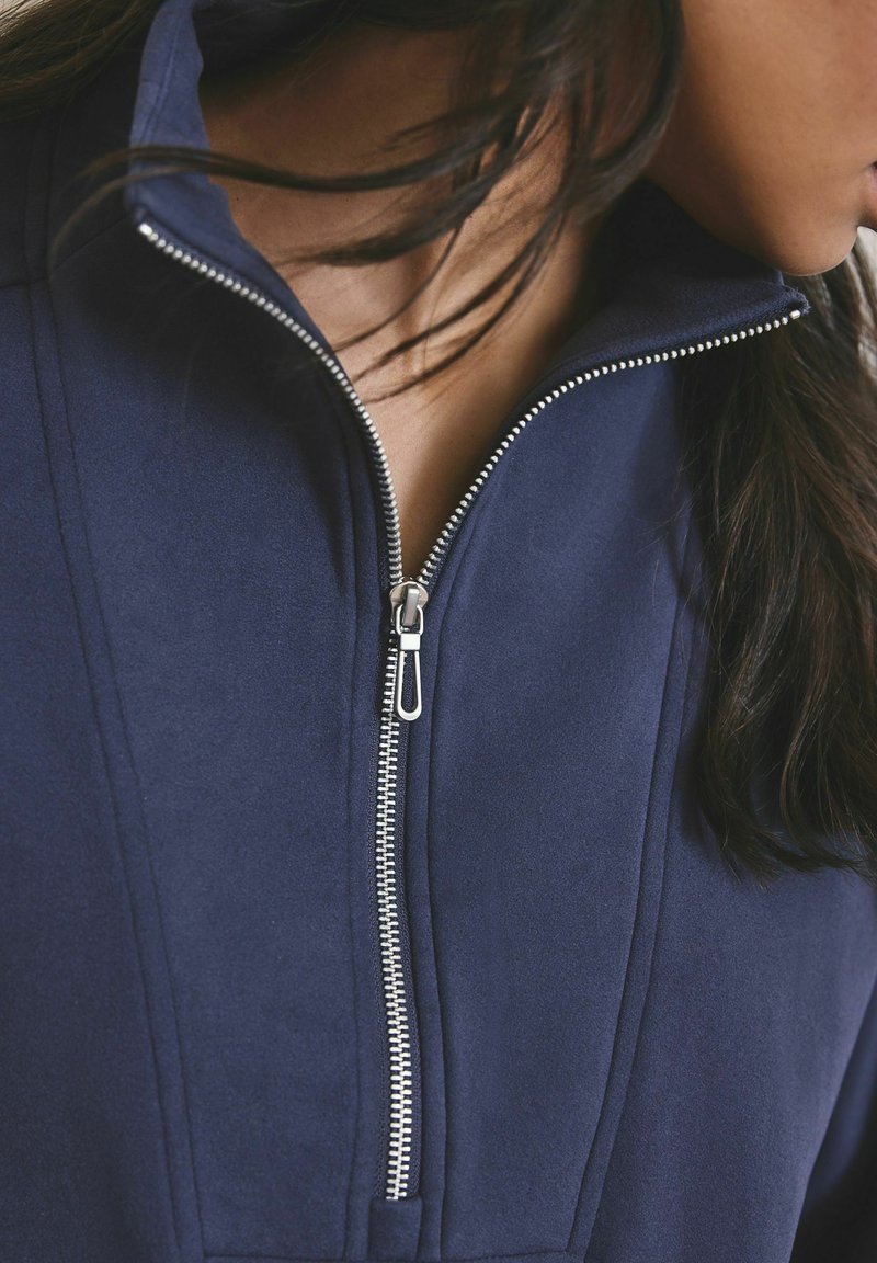 Felpa con zip blu navy con collo alto, cerniera argentata e texture liscia. Presenta dettagli di cuciture sulle spalle e sul petto.