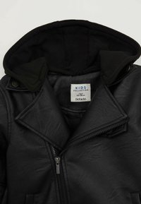 Veste pour enfants en simili cuir noir avec capuche noire amovible, devant zippé et col à revers. L'étiquette indique "COLLECTION ENFANTS" ainsi que les détails de taille.
