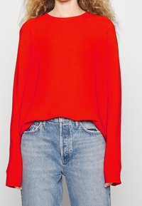 Rot gestrickter Pullover mit langen Ärmeln, lockerer Passform und weitem Ausschnitt, kombiniert mit hoch taillierten hellblauen Jeans.