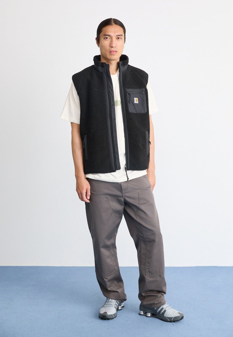 Schwarze Fleece-Weste mit Reißverschluss vorne, hohem Kragen und einer Brusttasche. Getragen über einem weißen T-Shirt und kombiniert mit grauen Hosen und Schuhen.