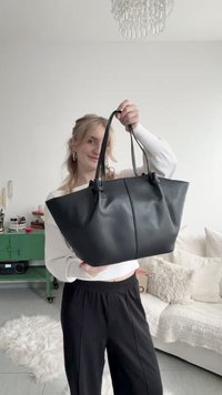 Žena u bijelom džemperu i crnim hlačama drži veliku crnu kožnu torbu u svijetlom dnevnom boravku s bijelom sofom.