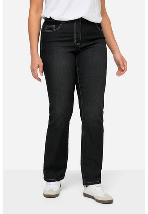 Persoon draagt zwarte, recht gesneden spijkerbroek met witte stiksels, witte sneakers en een wit overhemd tegen een effen achtergrond.