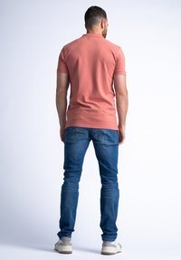 Chemise polo à manches courtes de couleur corail clair associée à un jean en denim bleu. La tenue présente une coupe décontractée et un tissu texturé. Des baskets viennent parfaire le look.