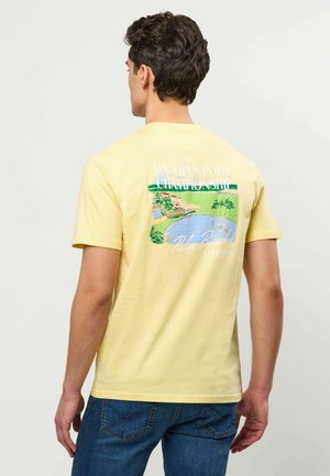 Mężczyzna w żółtej koszulce z kolorowym nadrukiem Palm Beaches Florida i U.S. Open Polo Championship na plecach, w połączeniu z niebieskimi dżinsami.