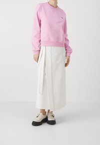 Růžová mikina s žebrovanými manžetami, spárovaná s bílou zavinovací sukní s bočními kapsami a kontrastním prošíváním. Doplňuje ji béžová obuv.