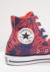 Zapatilla alta Converse All Star con patrón de teñido anudado rojo y azul marino, cordones blancos y parche de logo circular en el tobillo.