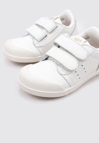 Zapatos de cuero blanco con un acabado suave, que cuentan con dos cierres de tira, punteras redondeadas y suelas de goma texturizadas. Acentos grises a lo largo de los lados.