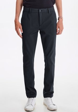 Dunkelgraue Hose aus gewobenem Stoff mit flachem Bund, Vorderaschen und slim-fit Design. Kombiniert mit weißen Sneakers.