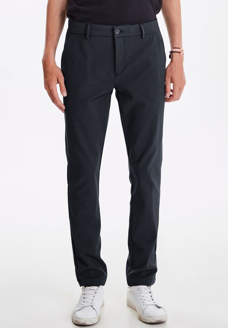Dunkelgraue Hose aus gewobenem Stoff mit flachem Bund, Vorderaschen und slim-fit Design. Kombiniert mit weißen Sneakers.