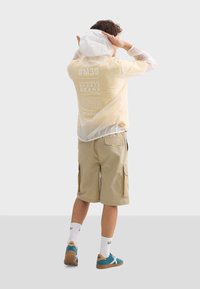 Coupe-vent léger et translucide avec capuche, teinte jaune, texte blanc au dos ; associé avec un short cargo beige et des baskets teal.