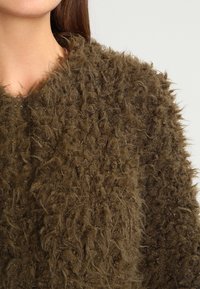 Brązowy, puszysty kardigan o teksturowanej strukturze z miękkim, niesfornym wyglądem. Posiada otwarty krój i minimalnie widoczne elementy metalowe.