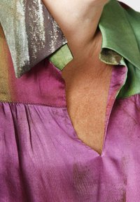 Bluse mit Kragen, die ein Farbverlauf von lila und grünen Schattierungen aufweist, mit einem strukturierten, gemusterten Kragen und Falten am Ausschnitt.