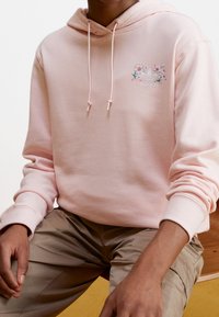 Sudadera con capucha de color rosa claro, confeccionada en tela suave, con un diseño floral y un logotipo de Adidas en el pecho. Cuerdas ajustables en el cuello.