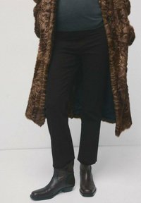 Osoba ubrana w czarne spodnie, ciemne skórzane botki za kostkę oraz długi, teksturowany brązowy futrzany płaszcz na ciemnej bluzce.