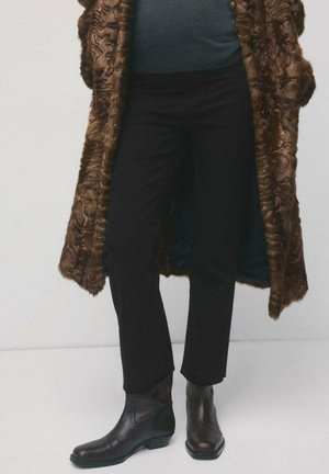 Persoon draagt zwarte broek, donkeren leren enkellaarzen en een lange, gestructureerde bruine bontjas over een donker shirt.