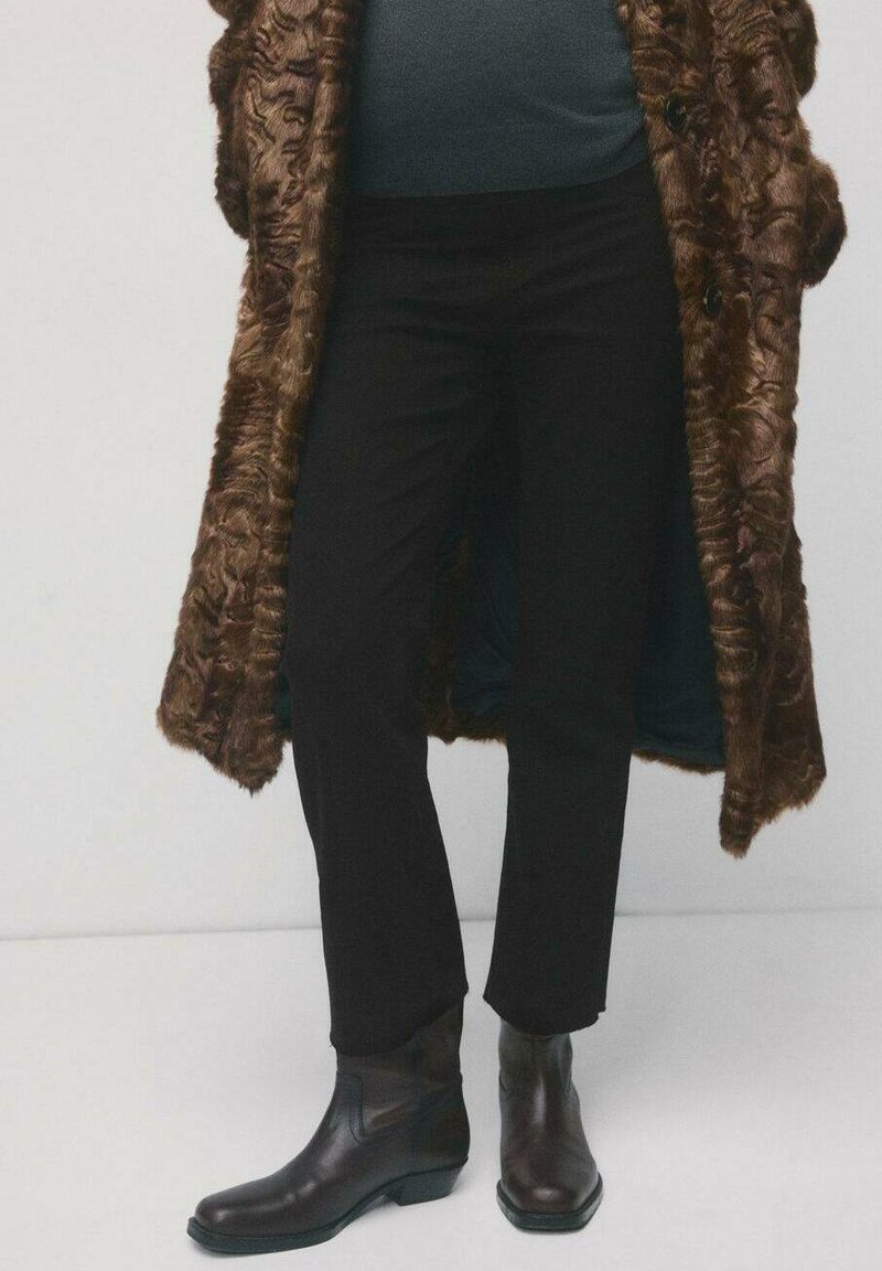 Osoba ubrana w czarne spodnie, ciemne skórzane botki za kostkę oraz długi, teksturowany brązowy futrzany płaszcz na ciemnej bluzce.