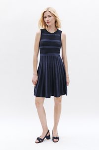 Ärmelloses marineblaues Kleid mit horizontalen blauen Streifen, ausgestelltem Rock und gerippter Textur. Getragen mit dunklen, klobigen Sandalen. Akzent mit einer goldenen Kettenhalskette.