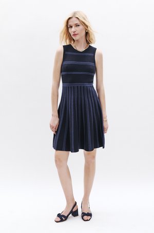 Robe sans manches bleu marine avec des rayures horizontales bleu, jupe évasée et texture côtelée. Portée avec des sandales à talons sombres. Accent en collier en chaîne dorée.