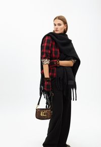 Una modella indossa uno scialle nero e un top a quadrati rossi con accenti leopardati, tenendo una borsa leopardata. Lo sfondo è bianco semplice.
