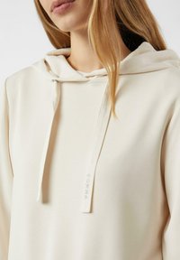 Beżowa bluza z kapturem ze ściągaczem, wykonana z miękkiego materiału; posiada subtelny detal tekstowy na sznurku. Gładka tekstura i luźny krój.