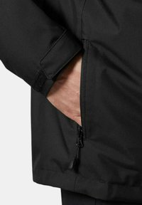 Chaqueta negra con un bolsillo con cremallera y puños de Velcro. El material es suave con una textura mate, diseñado para funcionalidad y comodidad.