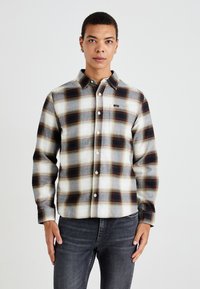 Camisa de franela de botones con un patrón de cuadros en blanco, gris y marrón oscuro. Cuenta con un cuello, mangas largas y un bolsillo en el pecho.