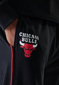 Svart hettegenser med et rødt og hvitt Chicago Bulls-logo på venstre lomme, med kontrastfargede røde detaljer på siden og ribbestrikkede mansjetter.