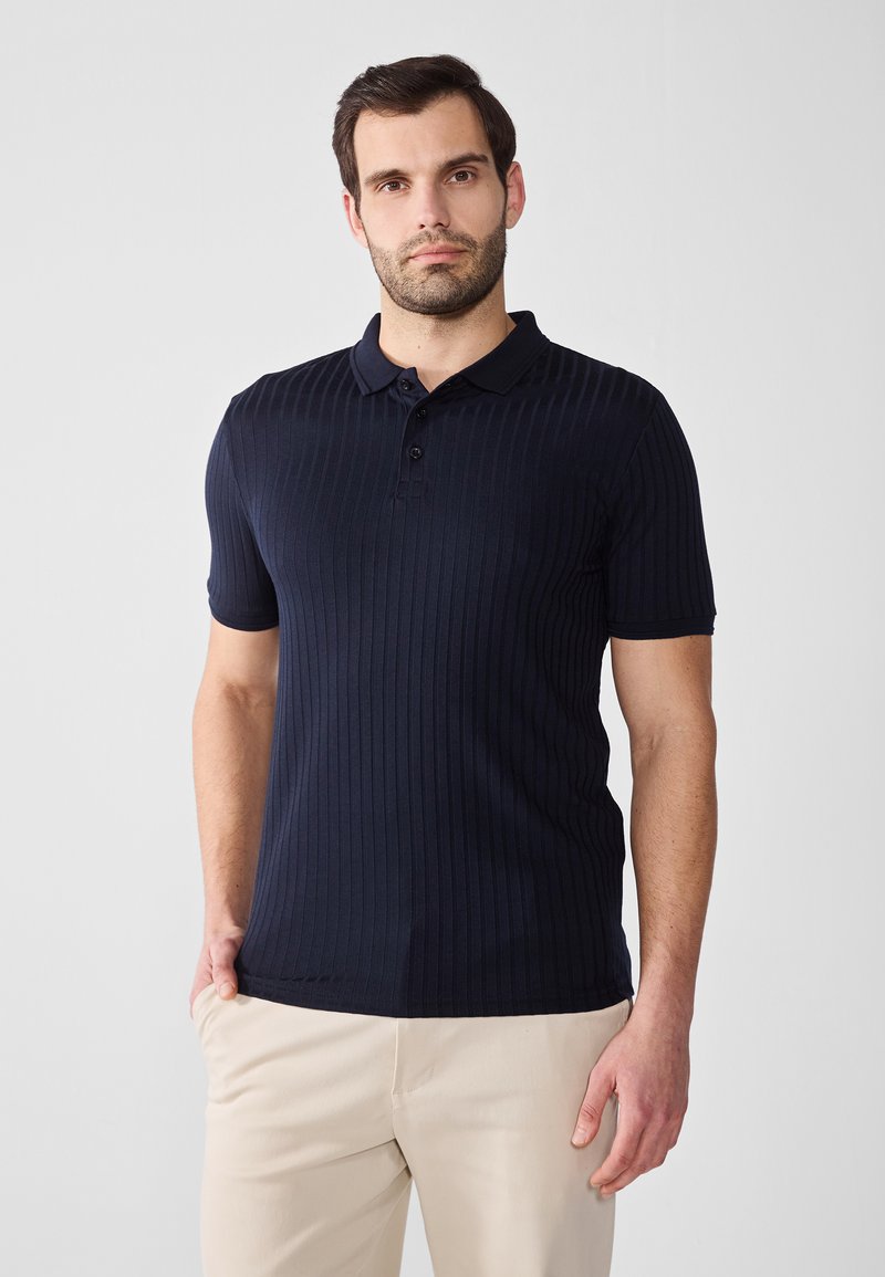 Polo de culoare albastru închis, cu dungi, cu mâneci scurte, guler cu trei nasturi și o textură moale. Asortat cu pantaloni bej, stând în fața unui fundal neutru.