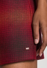 Un primo piano di una mano che tocca un tessuto a quadri rosso e nero con un piccolo cartellino logo sull'orlo, che mette in evidenza la texture e i dettagli del design.