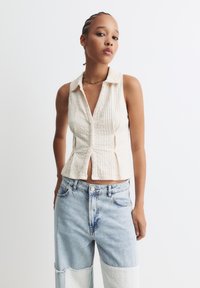 Crème ribbelgebreide mouwloze top met een kraag, knoopsluiting aan de voorkant en een getailleerd design, gecombineerd met lichtblauwe wijde jeans met patchwork.