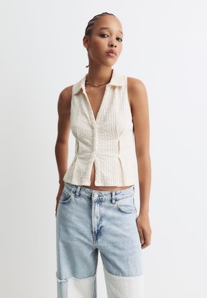 Cream ribbed knit sleeveless top with a collar, front buttons, and a fitted design, paired with light blue wide-leg jeans featuring patchwork.