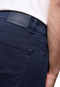 Pantalon en coton bleu marine au toucher lisse, avec une étiquette en cuir portant "Pierre Cardin", des poches latérales et des détails de couture visibles.