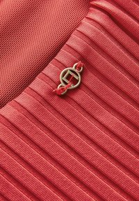 Rode stof met geplooide textuur, voorzien van een cirkelvormige gouden hardware accent en contrasterende rode stiksel details.