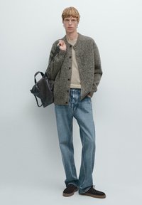 Brauner Strickcardigan mit Knöpfen, darunter ein beiges T-Shirt, blaue lässige Jeans und braune Wildlederschuhe; trägt eine dunkle Tasche.