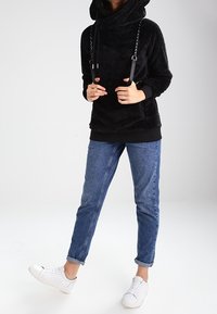 Henkilö, joka seisoo yhdellä jalalla ja jolla on päällään musta pörröinen huppari nyörityksillä, siniset käärityt farkut ja valkoiset lenkkarit.