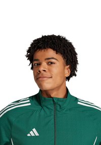Groene sportjack met een hoge kraag, rits aan de voorkant en drie witte strepen op elke schouder. De stof heeft een gestructureerde uitstraling.