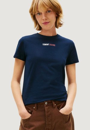 Mujer con cabello corto y rubio, vistiendo una camiseta azul marino de Tommy Jeans y pantalones marrones con las manos en los bolsillos, de pie frente a un fondo liso.