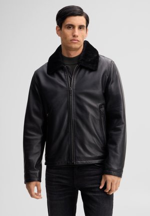 Mann trägt eine schwarze Lederjacke mit Pelzkragen, schwarze Hose und hat einen neutralen Gesichtsausdruck vor einem schlichten hellen Hintergrund.