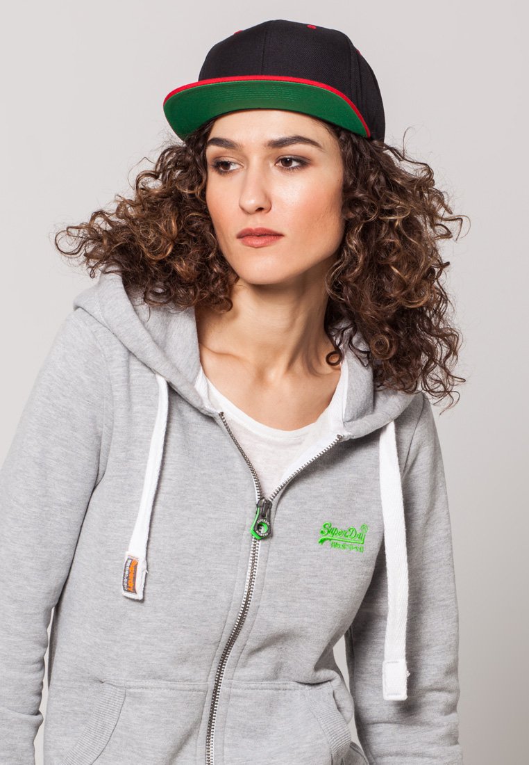 Felpa grigia con zip e coulisse bianche, abbinata a un cappellino nero con accento rosso sulla visiera, capelli ricci visibili.