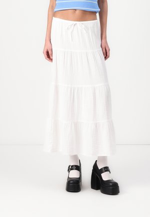 Jupe maxi blanche à étages en tissu léger avec une finition texturée, associée à des chaussures à plateforme noires et deschaussettes blanches.