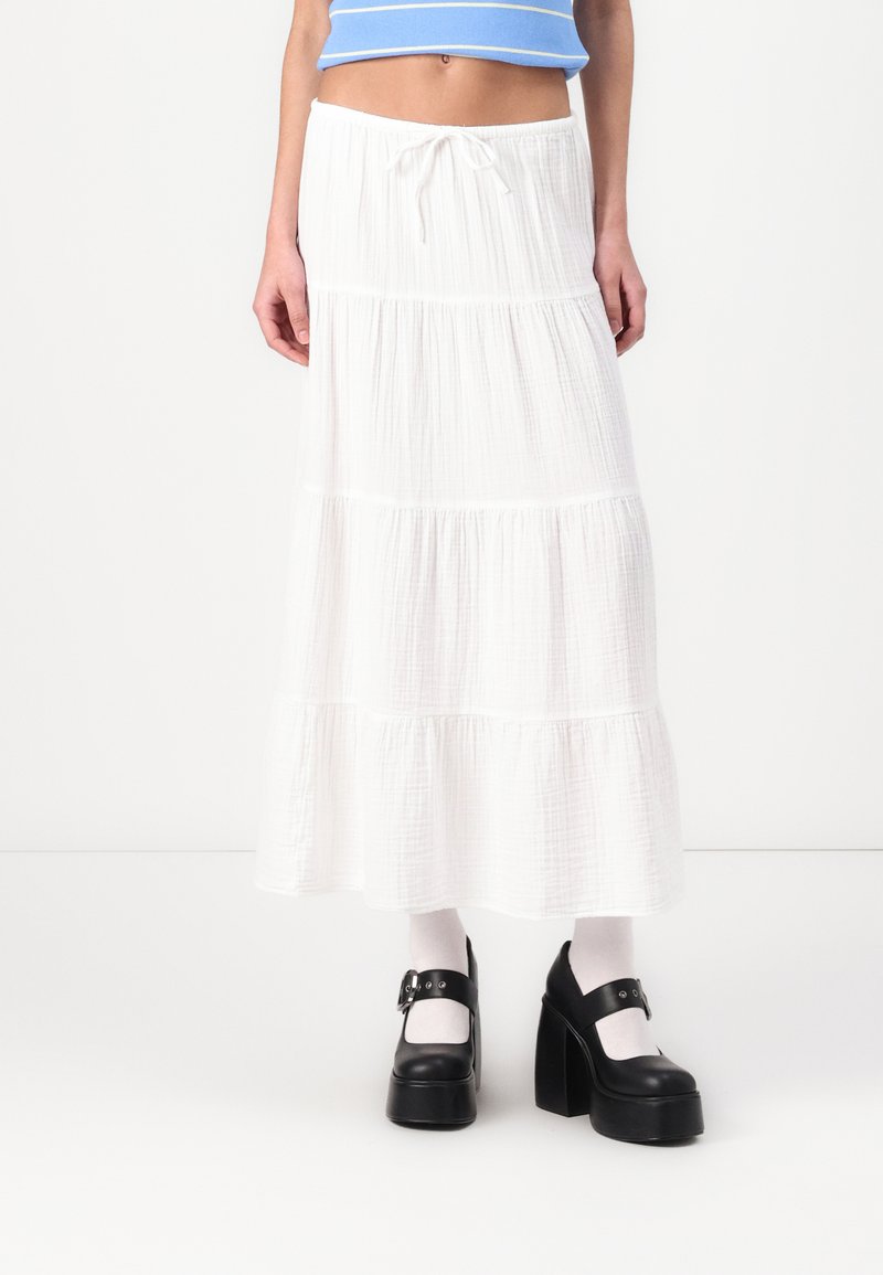 Falda maxi blanca en capas de tejido ligero con un acabado texturizado, combinada con zapatos de plataforma negros y calcetines blancos.
